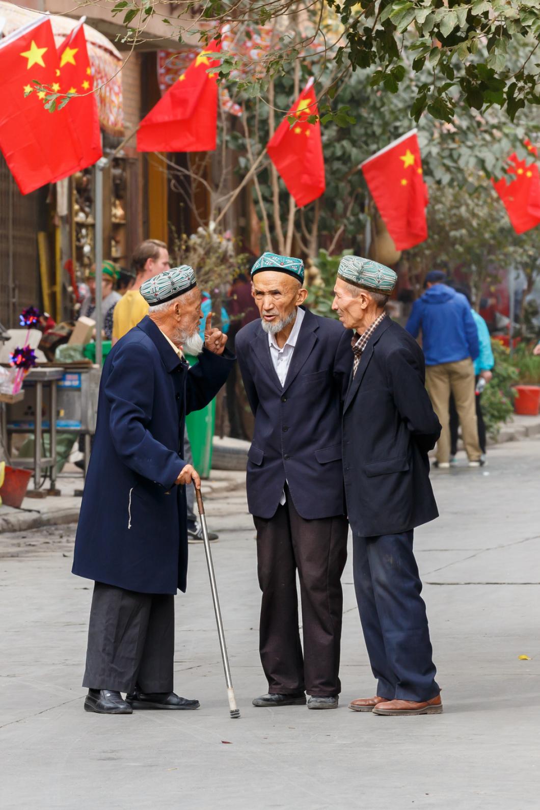 Drie oeigoerse oude mannen staan met elkaar te praten met chinese vlaggen op de achtergrond