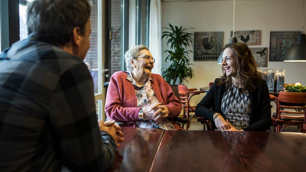 Een lachende oudere vrouw zit aan tafel met een jongere vrouw en een man