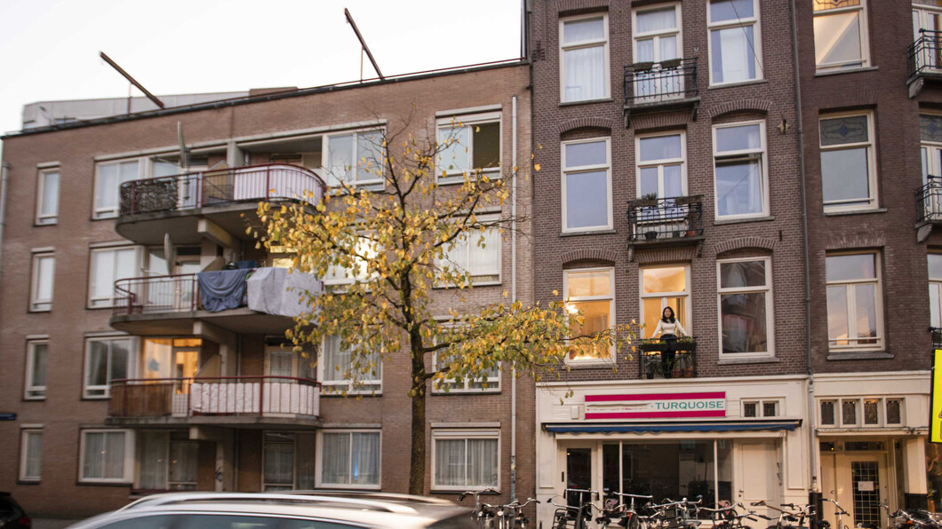 Het aangezicht van een aantal appartementen boven een buurtwinkel. Er staat iemand op het balkon.